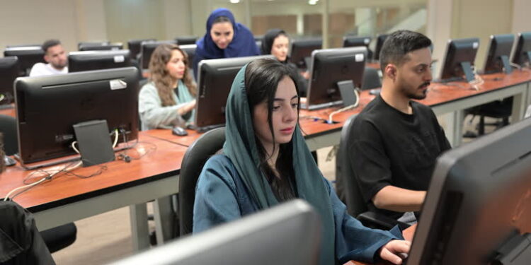 Young woman with scarf over head sitting next to man, using computers in technology suite in higher education institution and concentrating